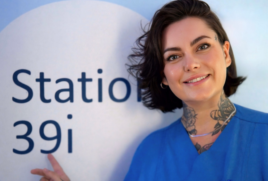 Charite Physican Assistant Victoria Hiebsch im Flur der Station 39i in die Kamera lächelnd und mit dem Finger auf das Stationsschild zeigend