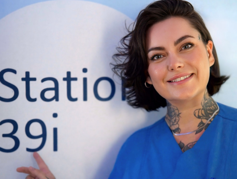 Charite Physican Assistant Victoria Hiebsch im Flur der Station 39i in die Kamera lächelnd und mit dem Finger auf das Stationsschild zeigend