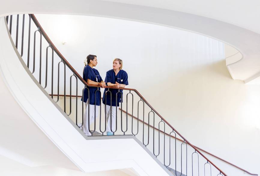Zwei Pflegekräfte stehen am Geländer einer Treppe und unterhalten sich