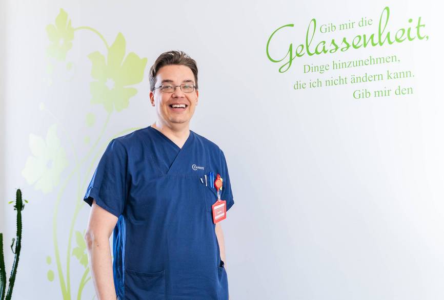 Pflegekraft Jan stehend im Büro in blauem Kasack.