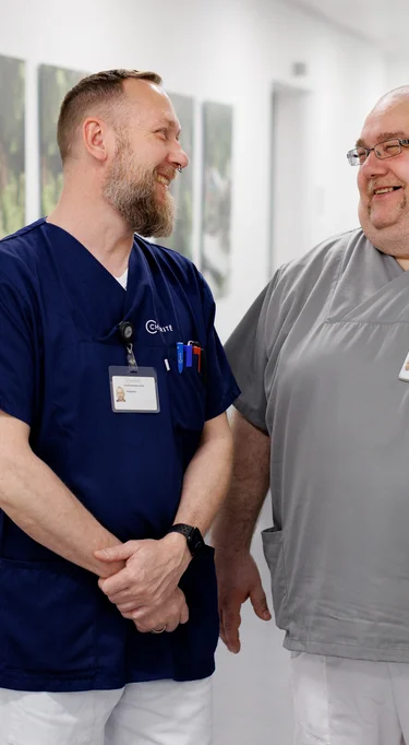 Eine Pflegefachkraft und eine Servicefachkraft stehen gemeinsam auf einem Stationsflur und unterhalten sich