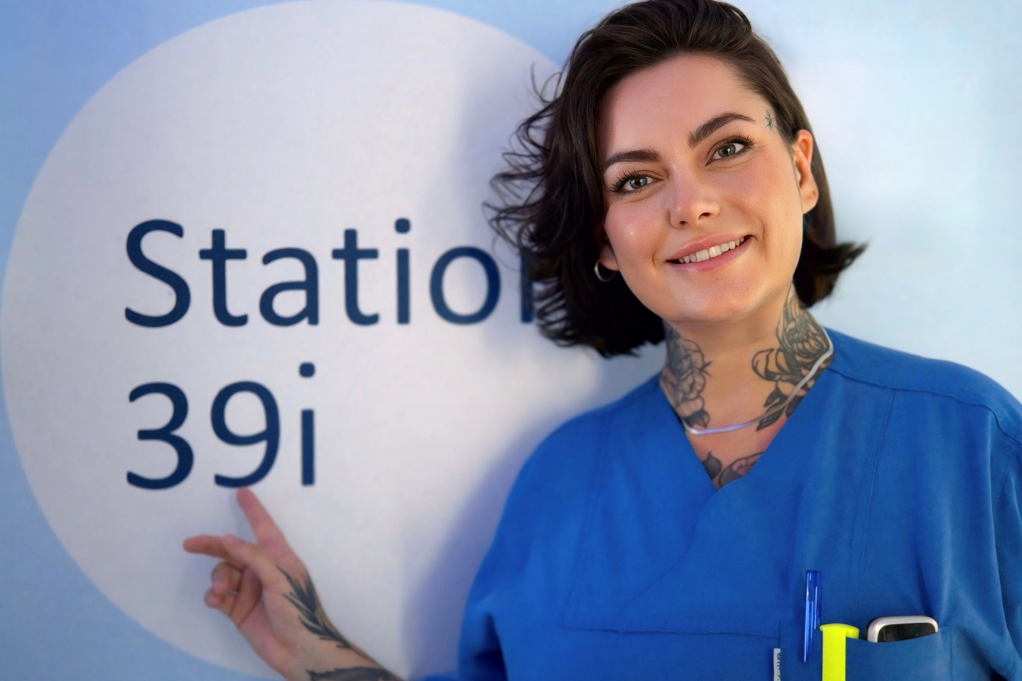 Charite Physican Assistant Victoria Hiebsch im Flur der Station 39i in die Kamera lächelnd und mit dem Finger auf das Stationsschild zeigend