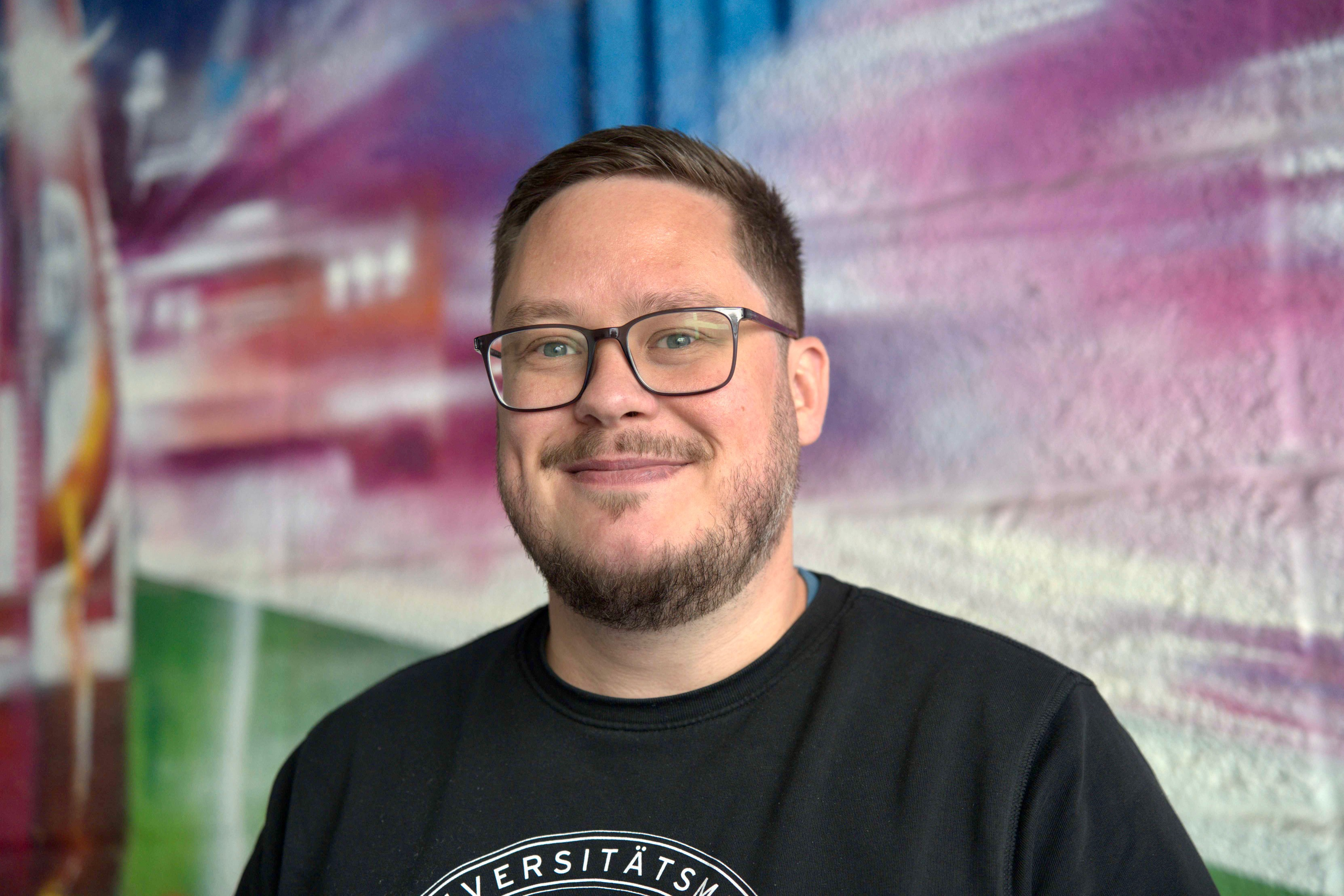 Charité Pflegefachkraft und Zentraler Praxisanleiter Pascal Hunger im Portrait vor einer bunten Grafitti-Wand