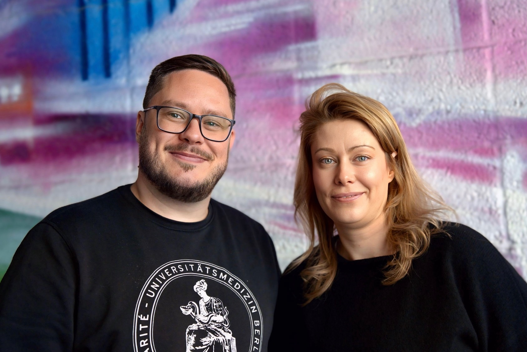 Charité Pflegefachkraft und Zentraler Praxisanleiter Pascal Hunger mit einer Master-Studentin vor einer bunten Graffiti-Wand im Portrait